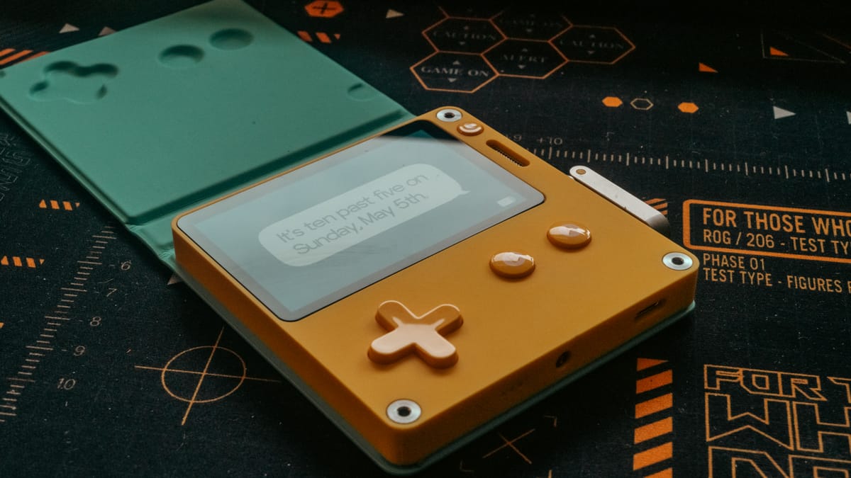 A yellow Playdate sits against a black and orange mat in a cyan colored case, still on its lock screen.
